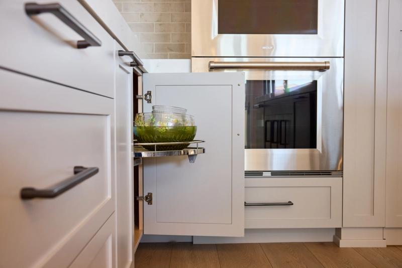 A close-up view of white shaker-style kitchen cabinetry with a pull-out cabinet door revealing a chrome shelf holding glass mixing bowls, positioned next to a stainless steel Wolf double oven.