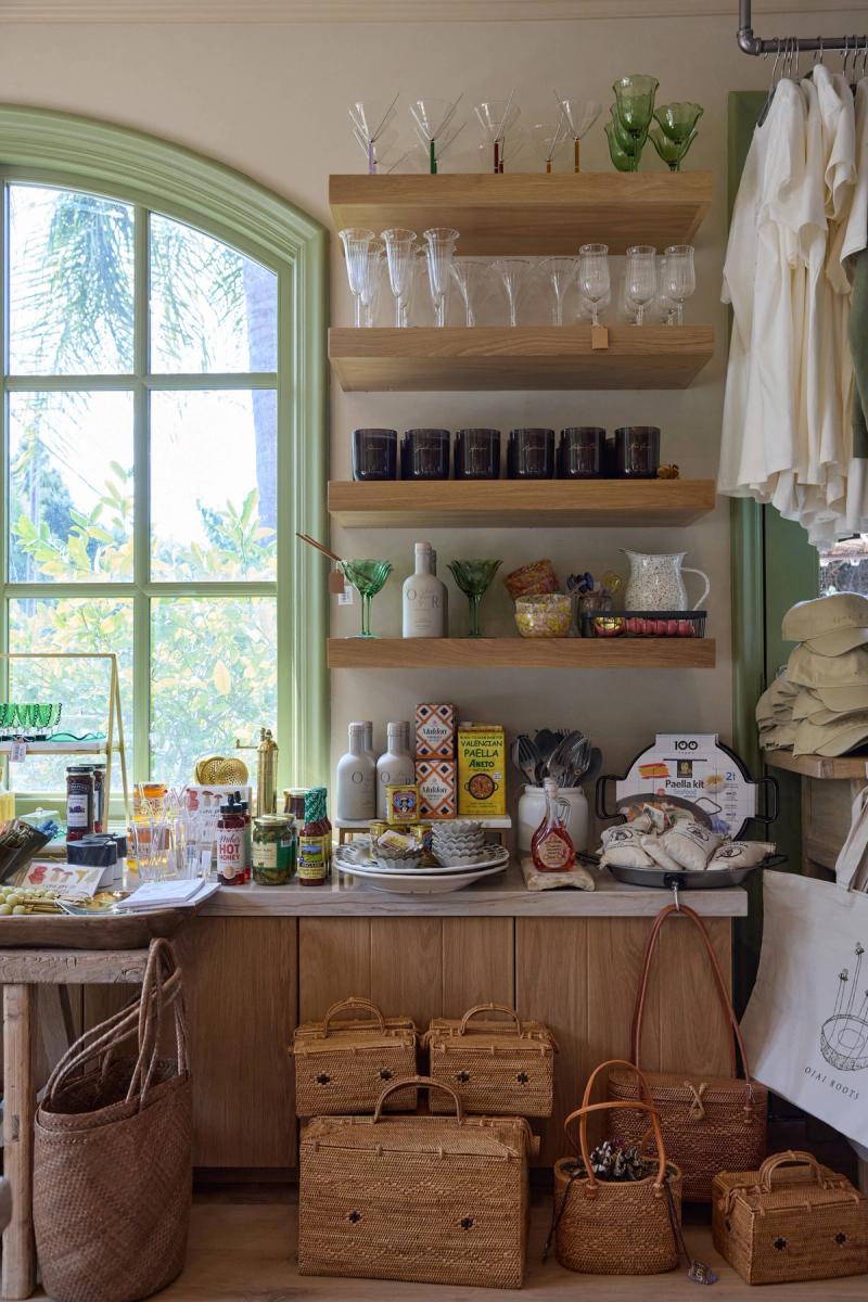 A charming retail or home shop interior featuring floating natural oak shelves mounted on a sage green wall beside an arched window, displaying glassware, candles, and gourmet food products, with woven baskets arranged on the floor below.