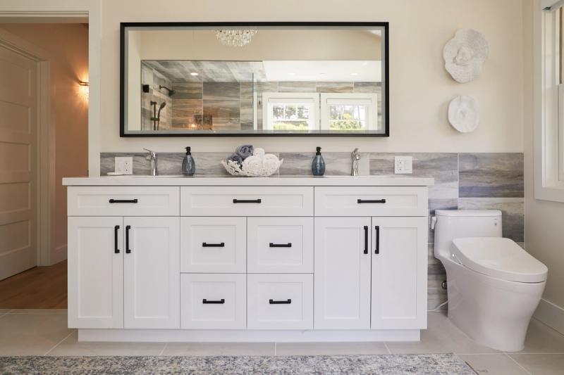 A bright master bathroom featuring a wide white shaker-style double vanity with black hardware, marble stone backsplash, large framed mirror, and a smart toilet, accented with coastal decor.