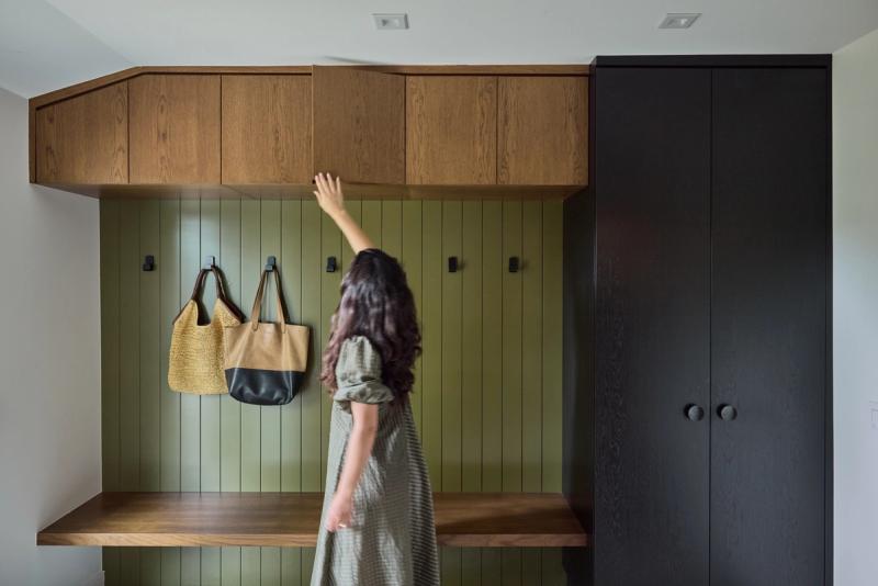 A woman reaches up to open an upper oak cabinet in a stylish mudroom featuring olive green vertical shiplap, black wall hooks with hanging bags, floating wood benches, and a dark floor-to-ceiling wardrobe cabinet.