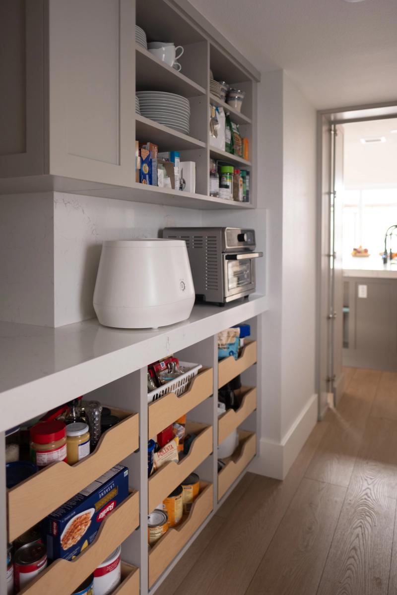 A well-organized butler's pantry featuring gray custom cabinetry with open upper shelves holding dishes and pantry items, a white quartz countertop with small appliances, and lower pull-out wooden drawers filled with dry goods and canned items.
