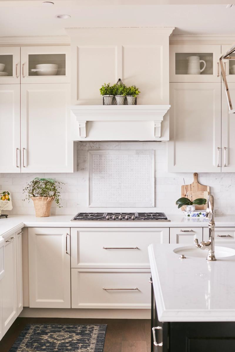 A bright, all-white traditional kitchen featuring custom inset cabinetry with polished nickel hardware, an elegant mantel-style range hood, marble backsplash, and white quartz countertops. Glass-front upper cabinets display white dishware and the space is accented with potted greenery.