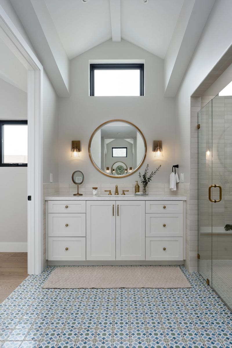 A bright, airy master bathroom featuring a white shaker-style vanity with brass hardware, a round gold-framed mirror, patterned blue and green floor tile, and a glass-enclosed shower. The vaulted ceiling with exposed beams and natural light from a clerestory window add architectural interest.