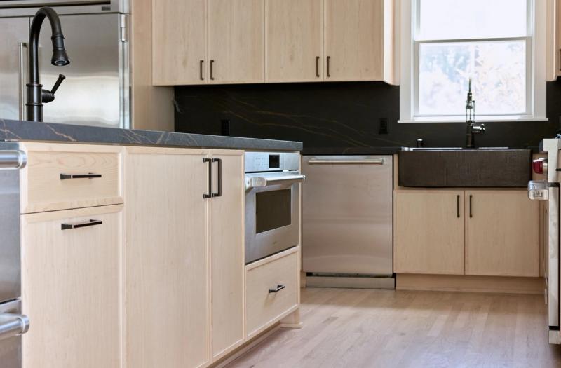A modern kitchen featuring light maple flat-panel cabinetry with matte black hardware, dark veined stone countertops and backsplash, stainless steel appliances, and a farmhouse-style sink near a bright window.
