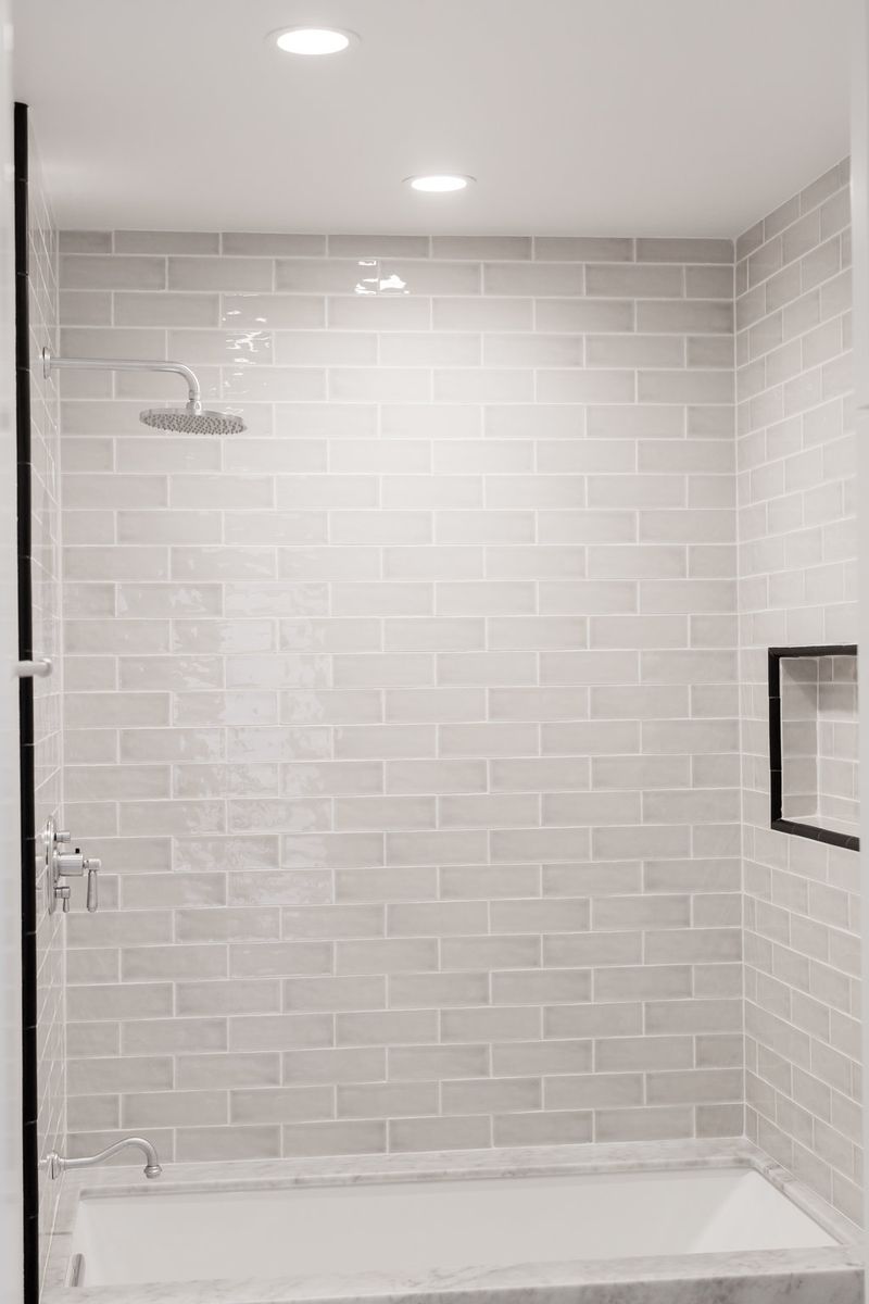 A clean, modern tub-shower combination featuring floor-to-ceiling light gray subway tile, a round rainfall showerhead, chrome fixtures, a built-in niche with black trim, and a white marble tub surround.