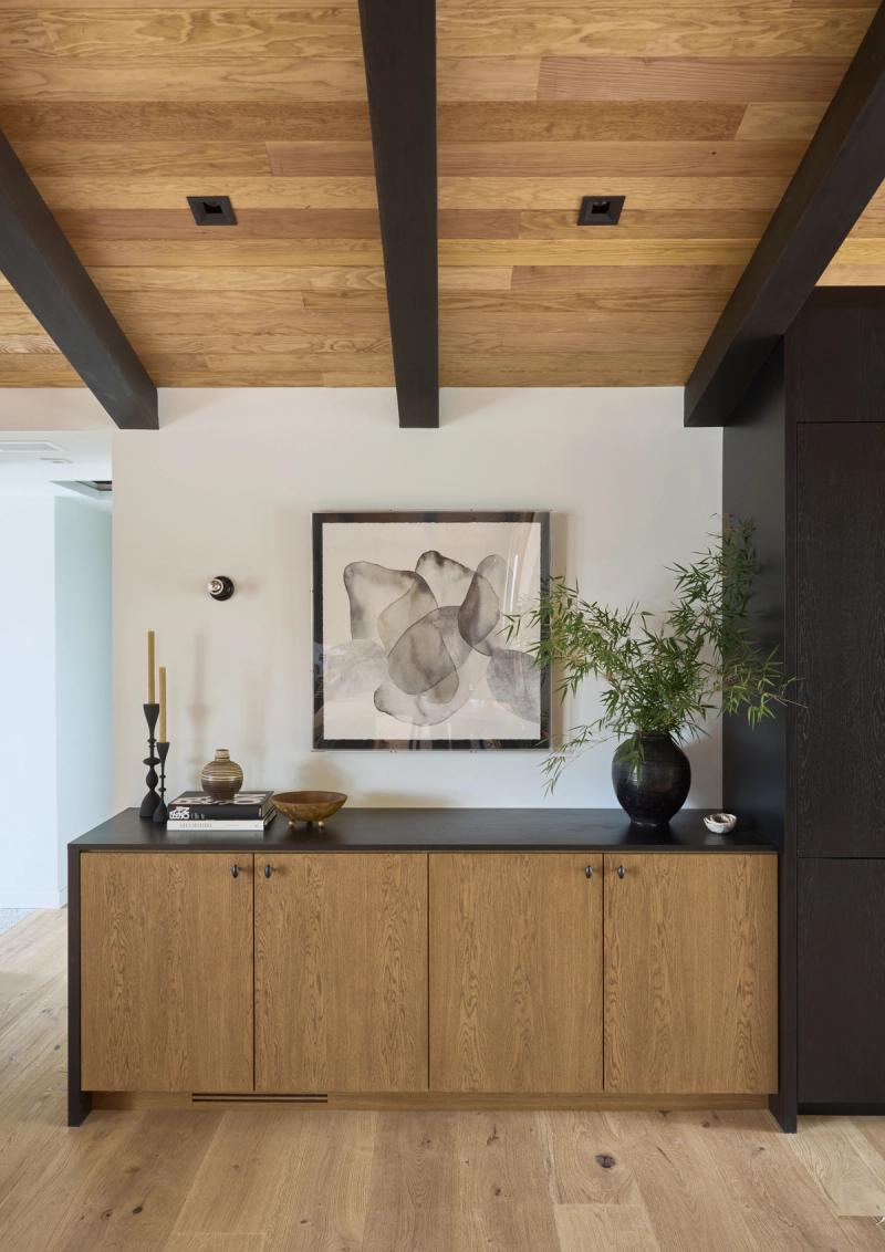 A custom wood credenza with warm oak cabinet doors and a dark matte top, styled with decorative objects and a bamboo plant, set beneath a wood-planked ceiling with dark exposed beams.