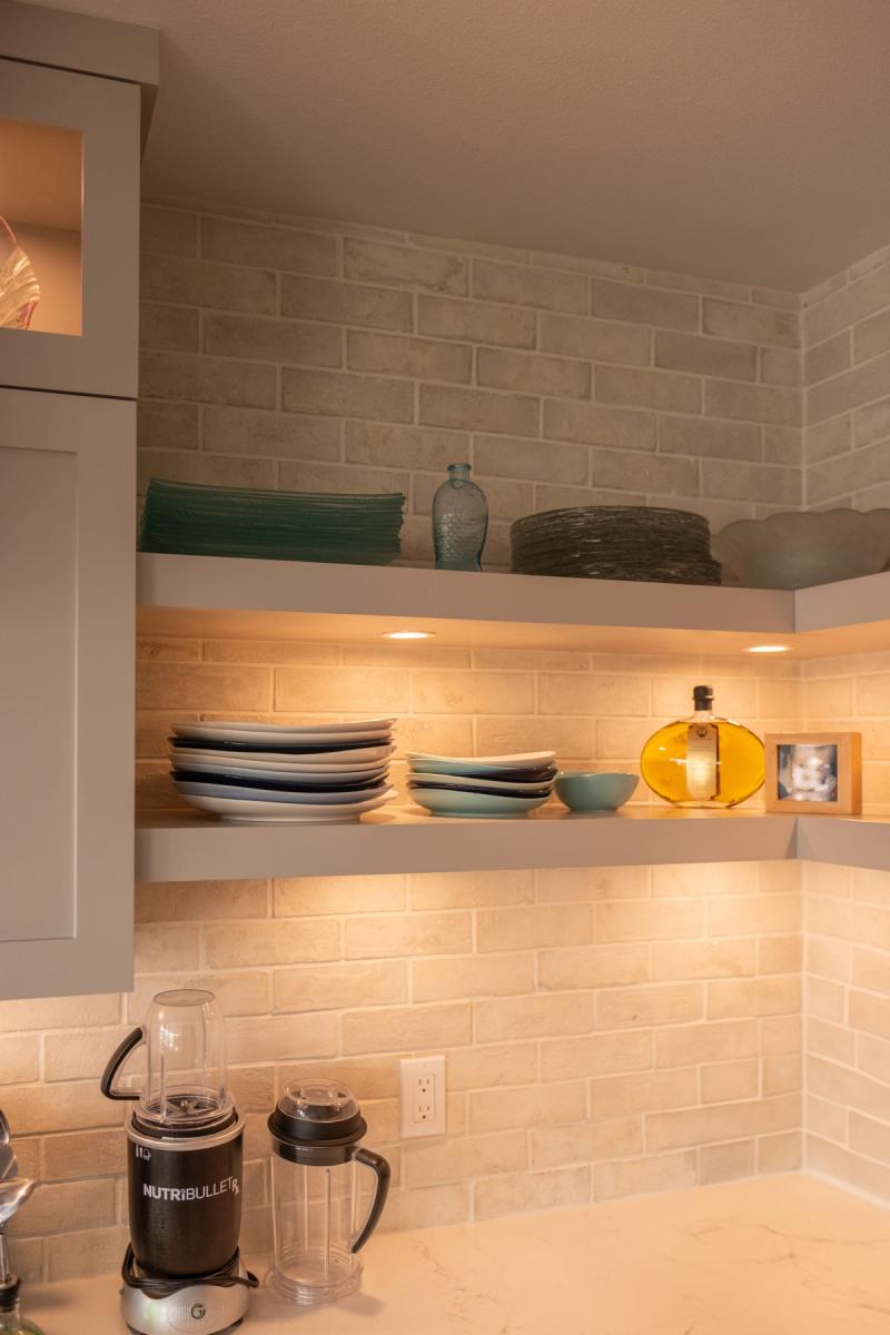 A warmly lit kitchen corner featuring custom floating open shelves with under-cabinet puck lights, displaying stacked plates, bowls, and decorative glassware against a cream brick tile backsplash, with a NutriBullet blender on the countertop below.