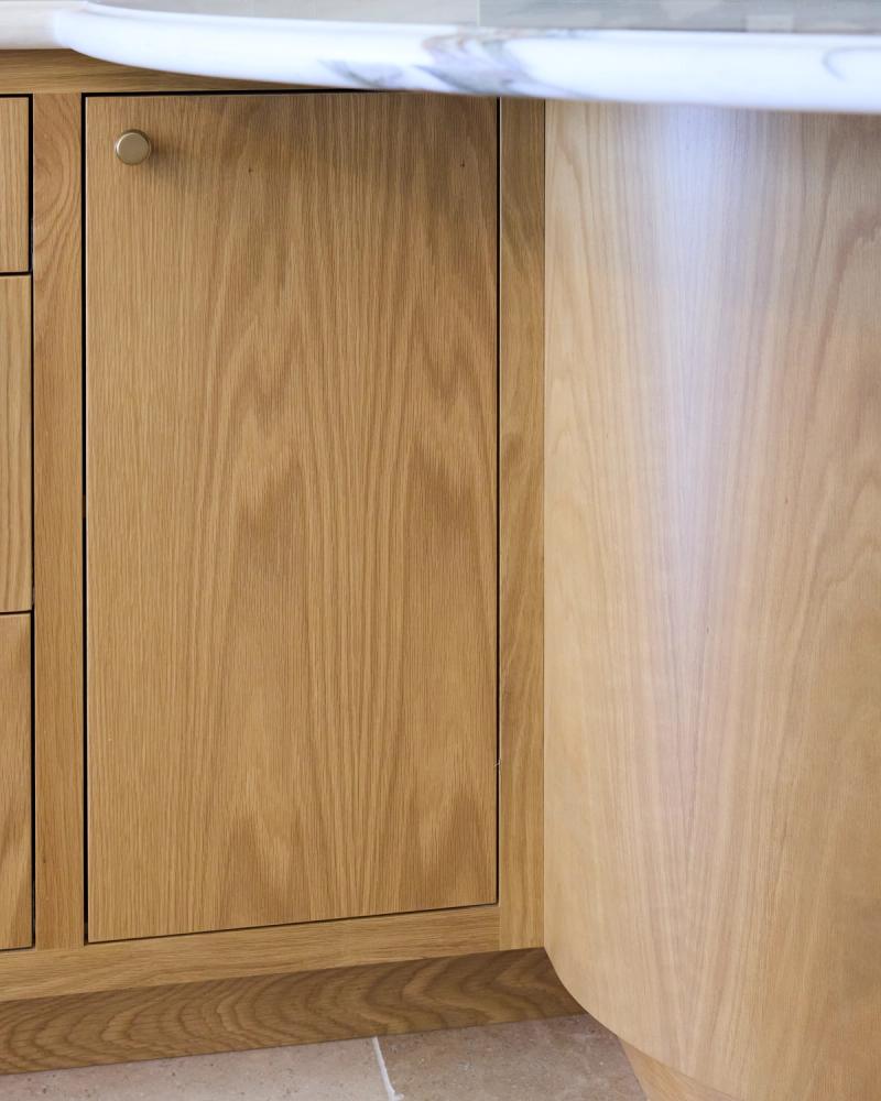 Close-up of custom natural oak kitchen base cabinets featuring flat-front doors with a brass knob hardware, paired with a white marble countertop and curved island end panel.