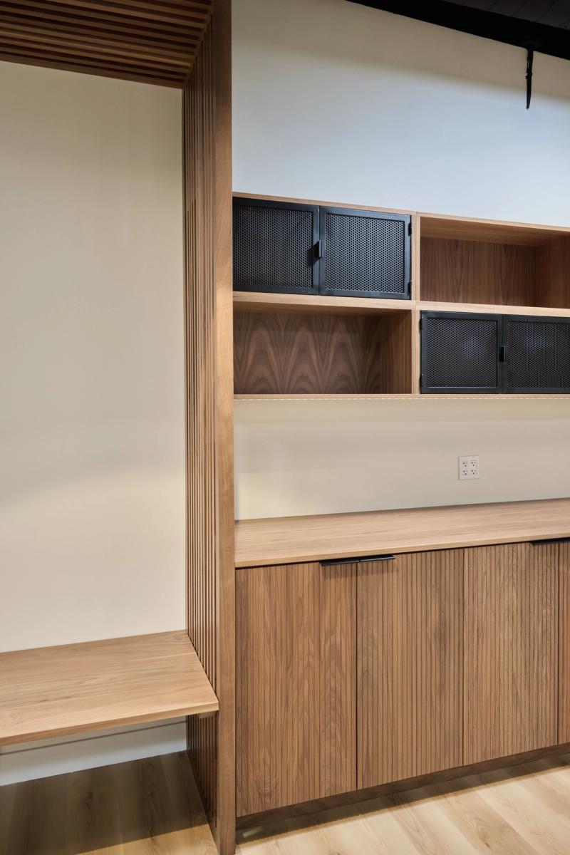 A custom walnut built-in unit featuring ribbed lower cabinet doors with black hardware, open shelving with perforated black metal cabinet doors on top, and a floating bench or desk surface extending to the left through a fluted wood portal.