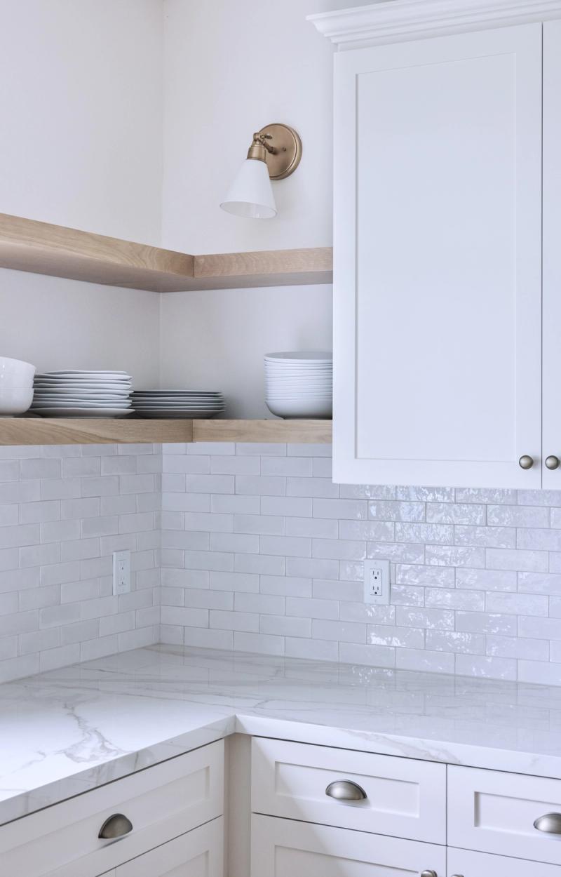 A bright, clean kitchen corner featuring white shaker cabinets, light wood open shelving stacked with white dishes, a white subway tile backsplash, marble countertops, and a brass wall sconce.