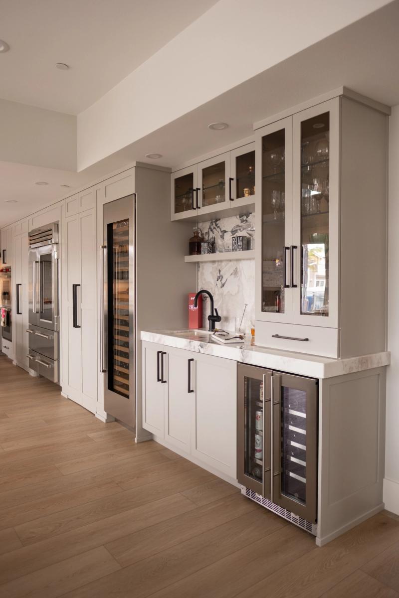 A custom wet bar area featuring gray shaker-style cabinetry with black hardware, glass-front upper cabinets displaying glassware, a marble backsplash, built-in wine cooler, and a tall wine column refrigerator, with stainless steel refrigerators visible in the background.