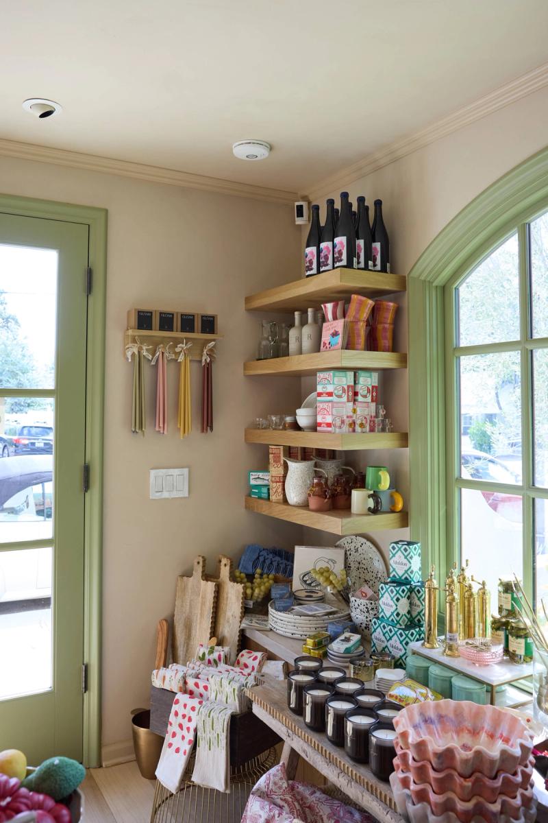 A charming retail or kitchen nook featuring natural oak floating corner shelves stocked with wine bottles, ceramics, and pantry goods, flanked by green-trimmed arched and French doors with colorful merchandise displayed on a wooden table below.
