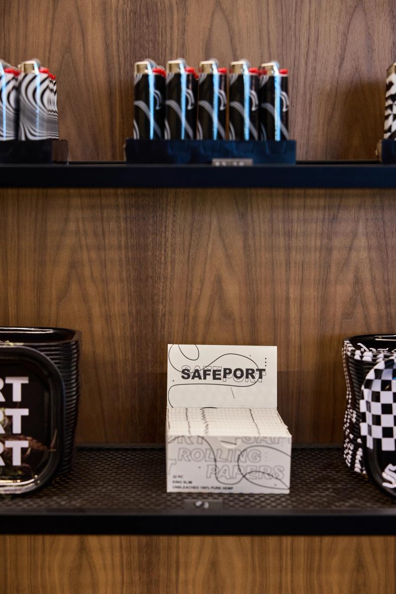 A close-up of a retail store shelf featuring custom dark-stained wood cabinetry with black metal shelving, displaying branded lighters, rolling papers, and decorative trays.