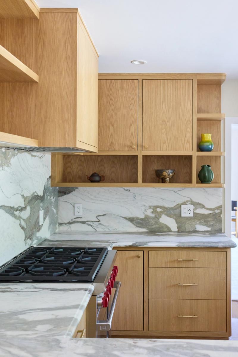 A modern kitchen featuring warm white oak cabinetry with brass hardware, open shelving displaying ceramic pottery, a dramatic white and green marble backsplash and countertops, and a professional gas range.