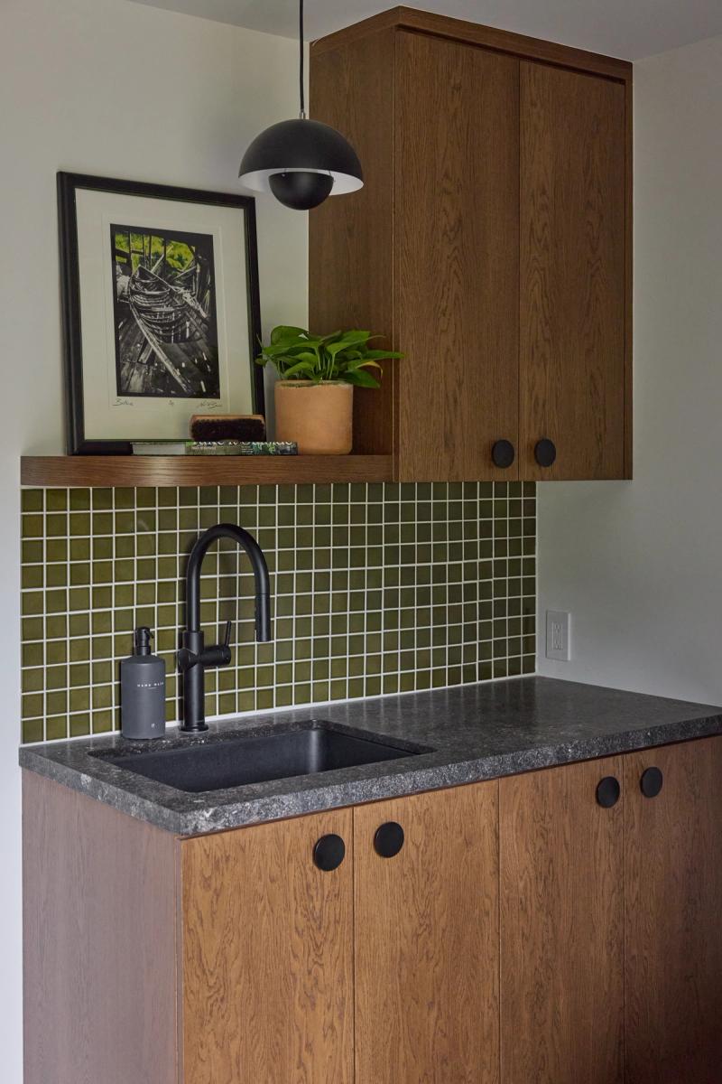 A compact wet bar or butler's pantry featuring warm oak cabinetry with matte black hardware, a dark granite countertop, black undermount sink, and a bold olive green mosaic tile backsplash. A floating wood shelf holds framed artwork and a potted plant above the counter.