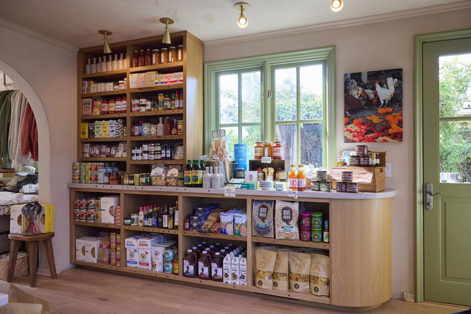 A bright, well-organized retail shop interior featuring custom light oak open shelving units and a curved display counter stocked with specialty food products, set against green-trimmed windows with natural light.