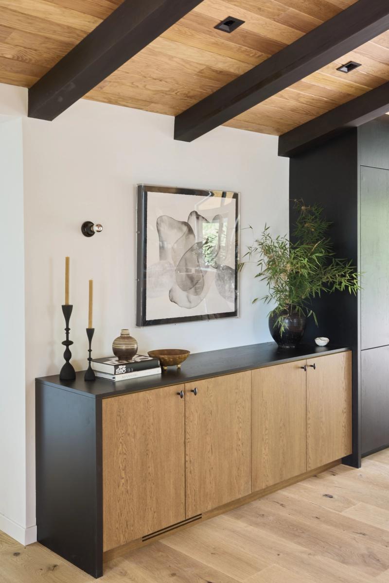A sleek entryway credenza with natural oak cabinet doors and a matte black top, styled with candle holders, books, a ceramic vase, and a large bamboo plant beneath an abstract framed artwork. The room features exposed black ceiling beams and a wood plank ceiling.