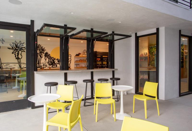 A bright modern outdoor patio area featuring yellow chairs, small white bistro tables, black bar stools, and folding pass-through service windows opening into a retail or café interior with colorful murals.