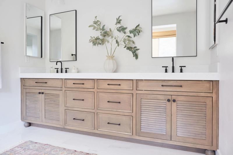 A wide double bathroom vanity featuring light natural wood cabinetry with louvered doors, black bar pulls, a white quartz countertop with two undermount sinks, matte black faucets, and two rectangular black-framed mirrors above.