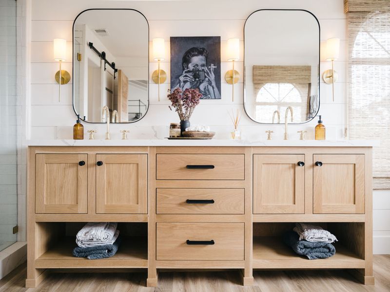 A wide double-sink bathroom vanity built from light natural oak wood, featuring black hardware, a white marble countertop, brass sconce lighting, two rounded rectangular mirrors, and open lower shelving with folded towels.