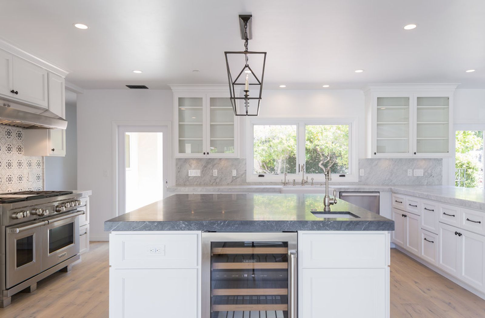A bright, spacious white shaker kitchen featuring a large center island with dark marble countertops, a built-in wine cooler, stainless steel Wolf range, glass-front upper cabinets, and a lantern pendant light.