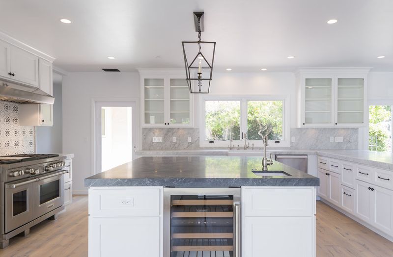 A bright, spacious white shaker kitchen featuring a large center island with dark marble countertops, a built-in wine cooler, stainless steel Wolf range, glass-front upper cabinets, and a lantern pendant light.