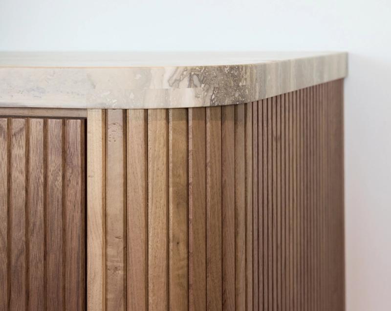 A close-up detail shot of a custom wood cabinet featuring vertical fluted panel doors in a warm walnut tone, topped with a travertine or marble stone surface with natural veining.