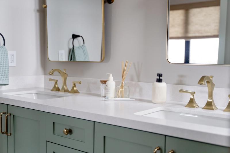 A double bathroom vanity with sage green shaker-style cabinets, white quartz countertop, brushed brass faucets and hardware, and two gold-framed mirrors. The counter is styled with a reed diffuser, soap dispensers, and a toothbrush.