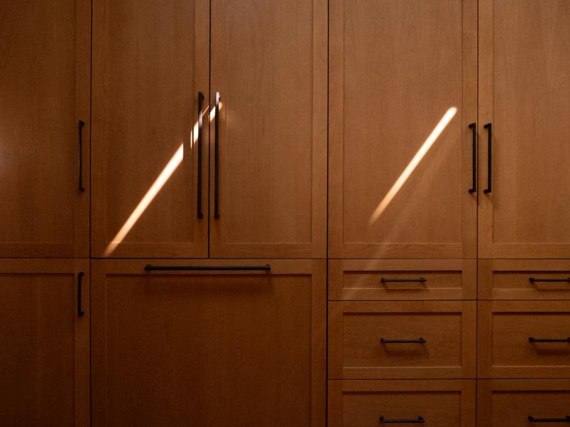 A floor-to-ceiling wall of custom wood cabinetry in a warm honey oak finish, featuring black bar pulls and diagonal rays of natural sunlight across the cabinet doors.