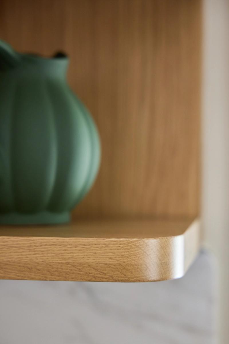 A close-up detail shot of a rounded-edge natural oak shelf inside a built-in cabinet, with a matte green ribbed ceramic vase resting on it.