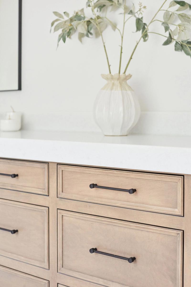 A close-up of a custom light wood bathroom vanity with raised panel drawers and matte black bar pulls, topped with a white quartz countertop and decorated with a ribbed ceramic vase holding eucalyptus branches.