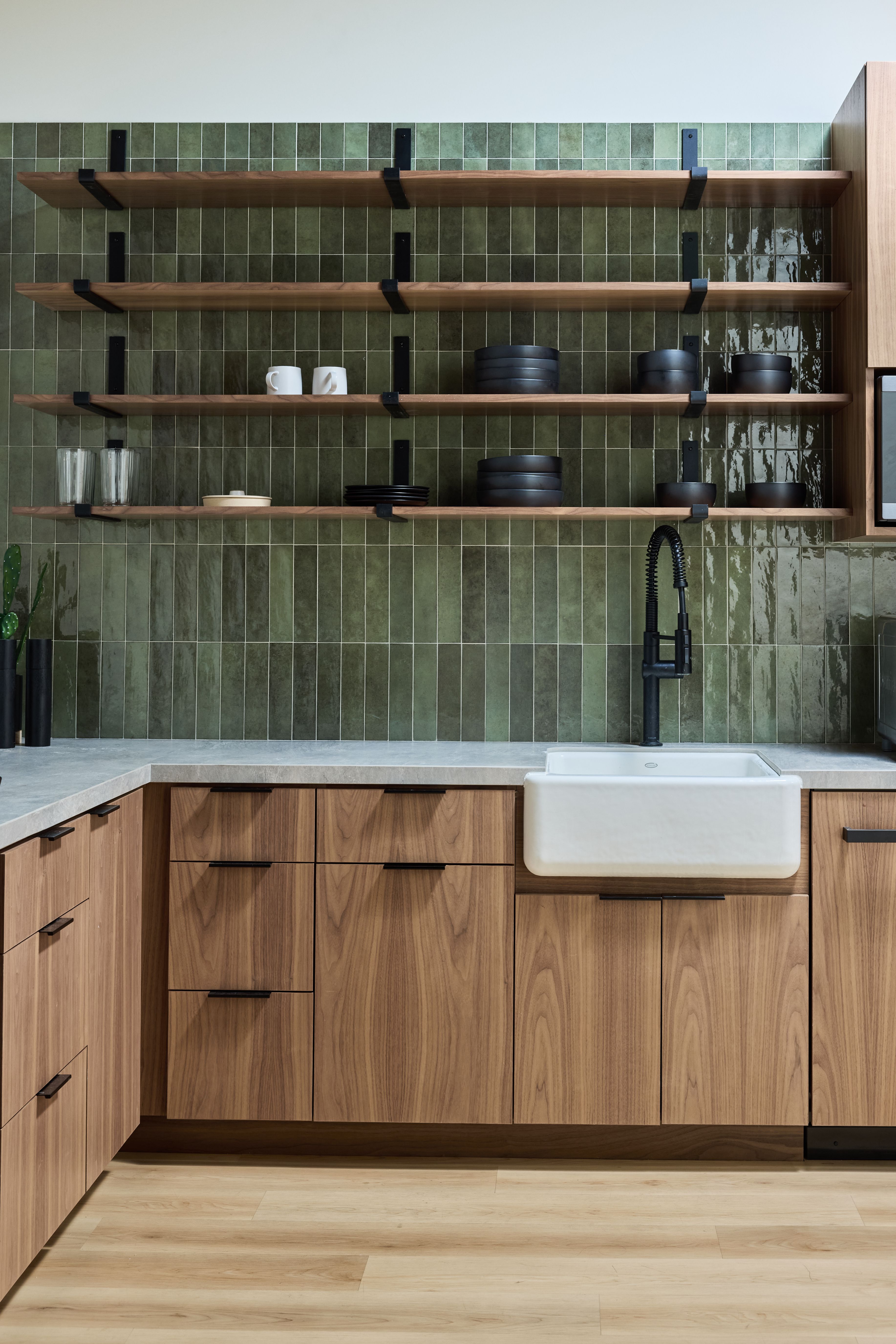 A modern kitchen featuring flat-front walnut cabinets with black hardware, a white farmhouse sink, matte black faucet, gray stone countertops, and floor-to-ceiling green glazed tile backsplash with open wood shelving displaying black dishware.