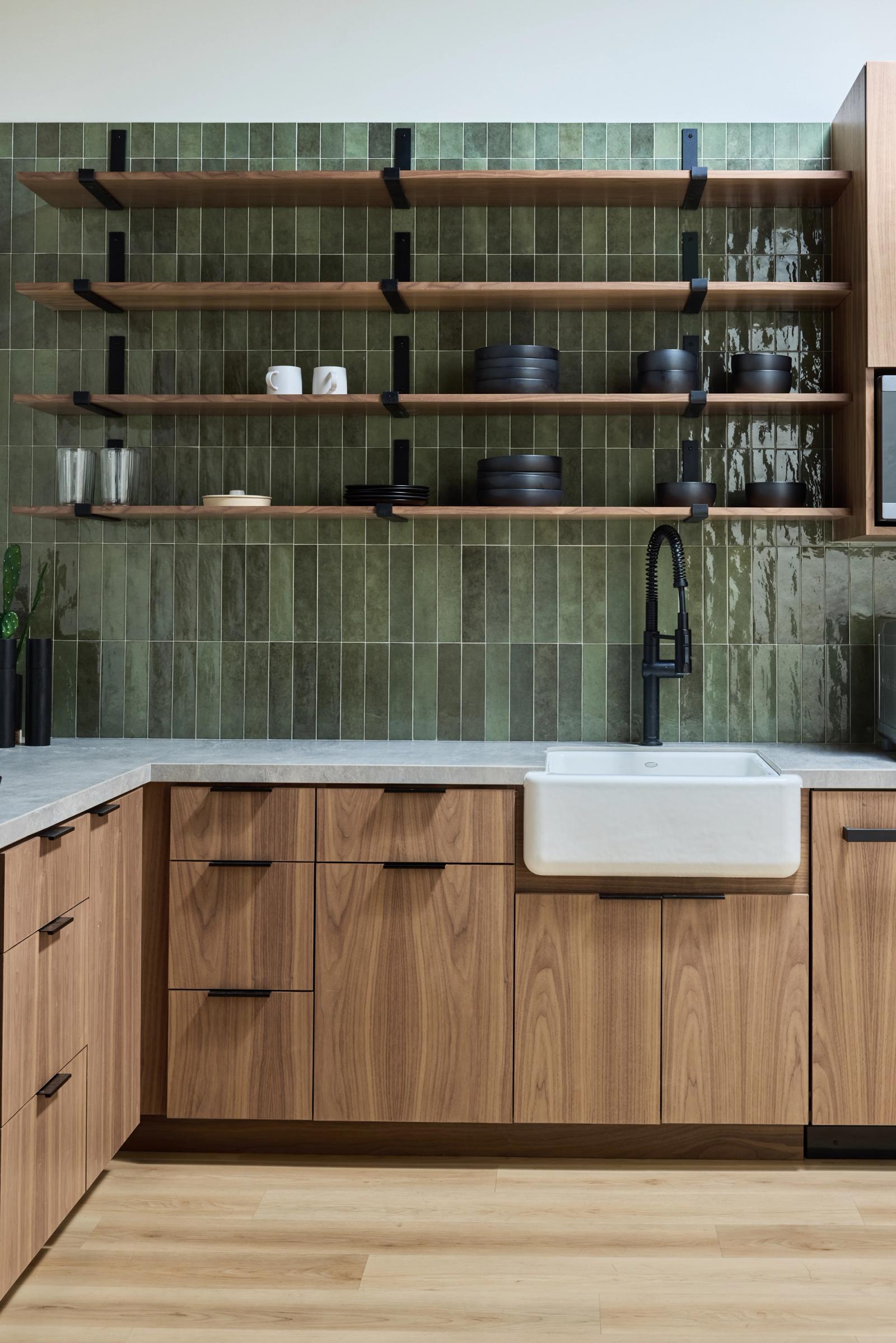 A modern kitchen featuring flat-front walnut cabinets with black hardware, a white farmhouse sink, matte black faucet, gray stone countertops, and floor-to-ceiling green glazed tile backsplash with open wood shelving displaying black dishware.