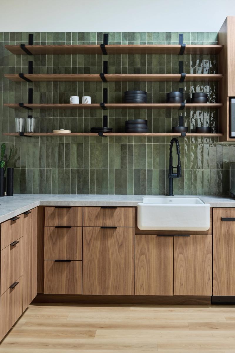 A modern kitchen featuring flat-front walnut cabinets with black hardware, a white farmhouse sink, matte black faucet, gray stone countertops, and floor-to-ceiling green glazed tile backsplash with open wood shelving displaying black dishware.