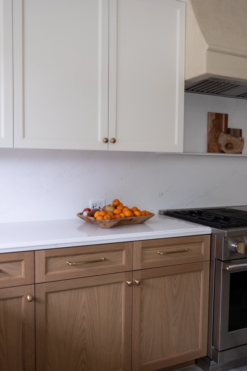 A custom kitchen featuring white upper cabinets with brass knobs and natural oak lower cabinets with brass pulls, white quartz countertops and backsplash, a stainless steel gas range, and a wooden bowl of oranges on the counter.