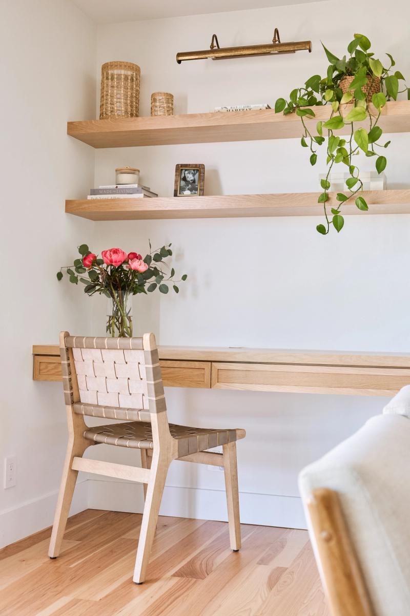A light wood floating desk with built-in drawer and matching open shelves mounted on a white wall, styled with a woven leather chair, pink roses, books, and a trailing pothos plant.