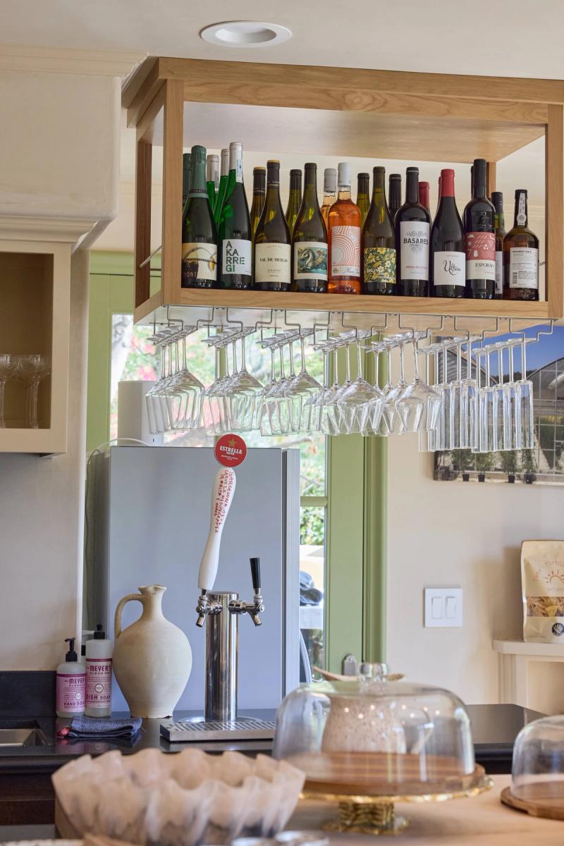 A custom white oak floating shelf above a bar area holds a collection of wine bottles, with hanging stemware racks below and a draft beer tap on the counter.