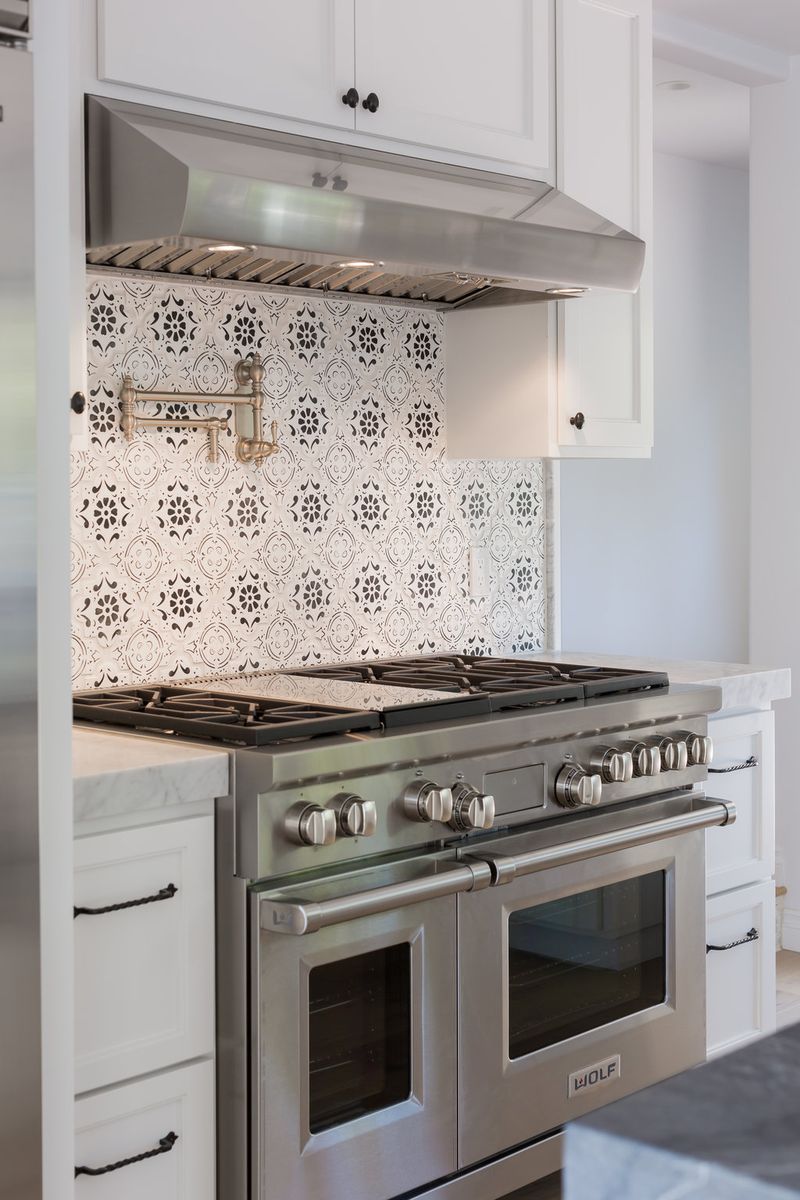 A professional kitchen featuring white raised-panel cabinetry, a stainless steel Wolf dual-fuel range with six burners and double ovens, a decorative black and white patterned tile backsplash, and a stainless steel range hood with a brass pot filler.