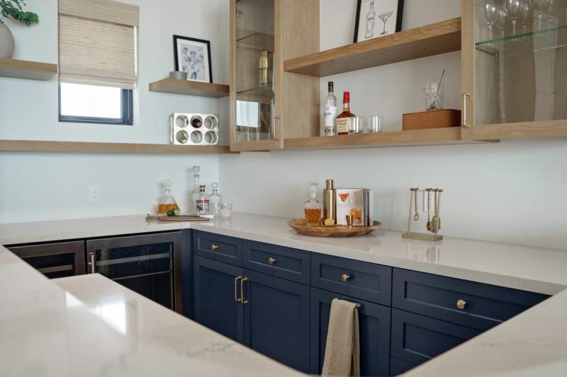 A stylish home wet bar featuring navy blue shaker-style cabinets with brass hardware, white quartz countertops, and open natural oak shelving stocked with spirits and glassware.