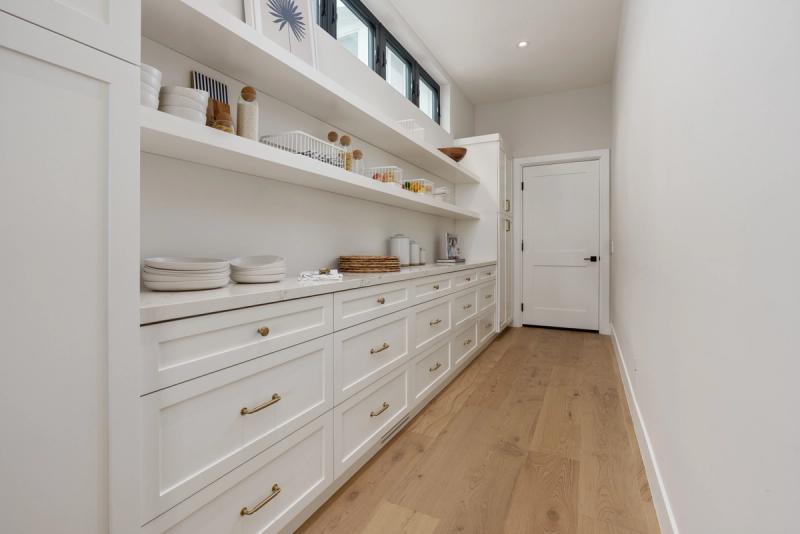 A bright, spacious butler's pantry featuring floor-to-ceiling white shaker cabinets and drawers with brass pulls, open shelving stocked with dishes and pantry items, a white quartz countertop, light oak hardwood floors, and a row of clerestory windows near the ceiling.