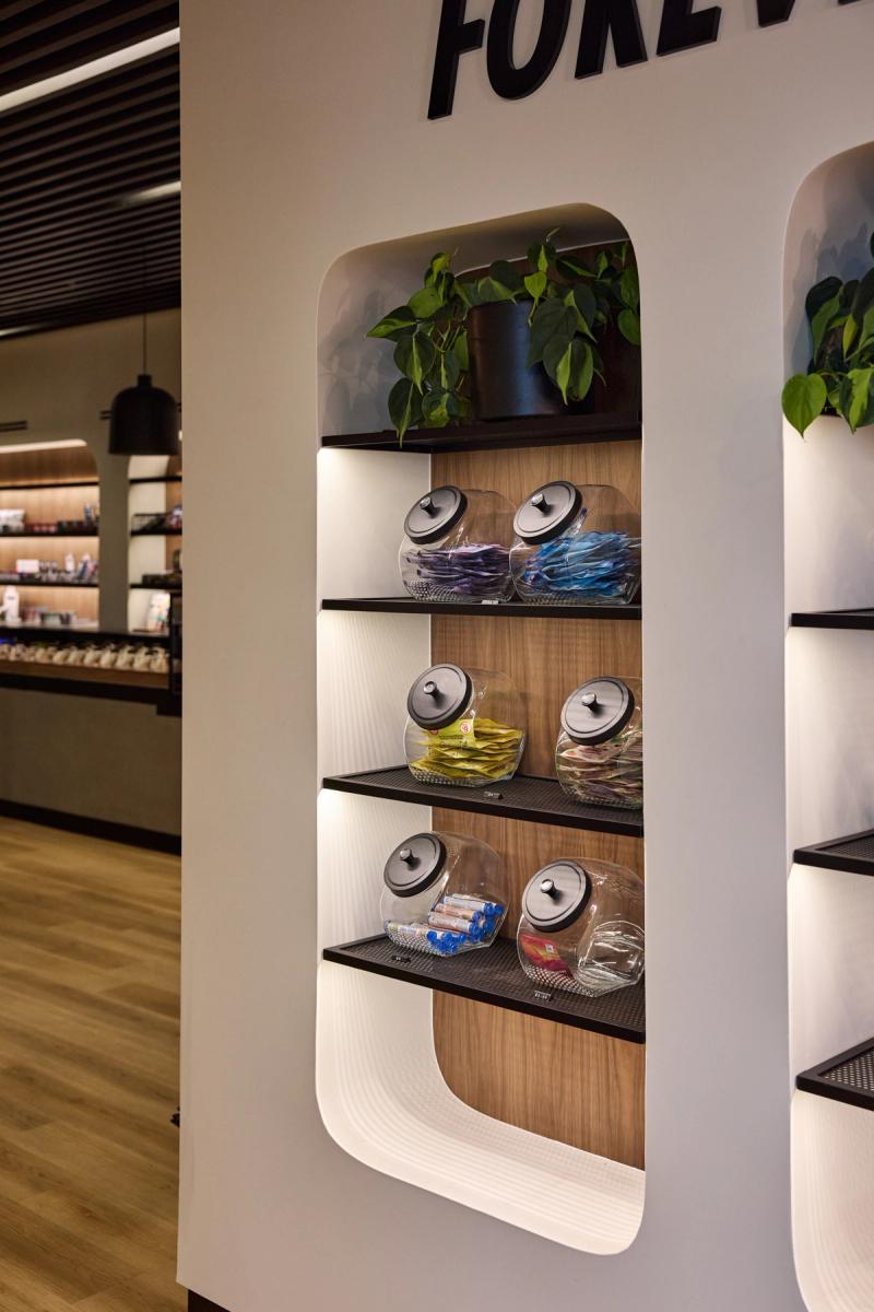 A modern built-in display wall unit with rounded arch niches, walnut wood backing, black mesh shelving, and glass jars filled with colorful packaged items, topped with green potted plants.
