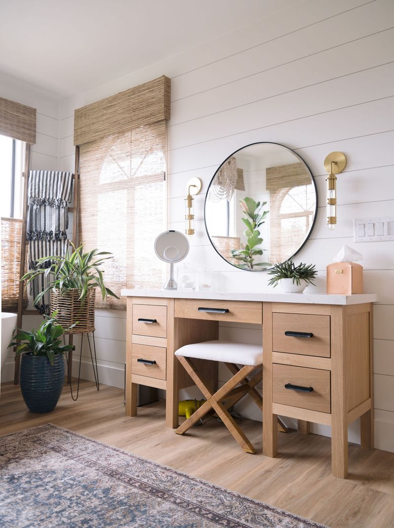 A bright, airy bathroom vanity area featuring a light oak wood vanity with black hardware, white quartz countertop, round mirror with black frame, and gold wall sconces mounted on white shiplap. A padded X-bench stool sits in the knee space.