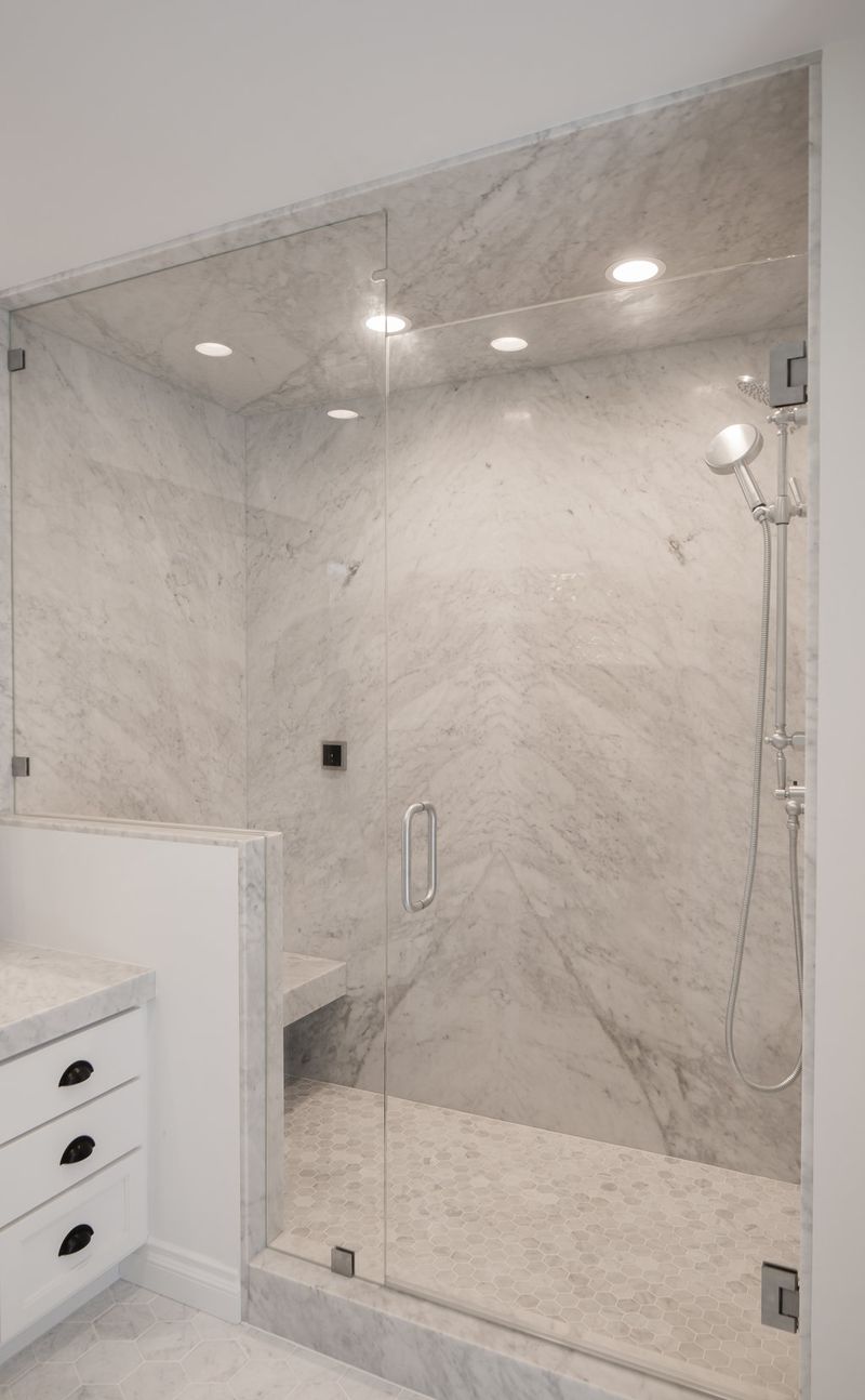A luxurious walk-in shower enclosure featuring floor-to-ceiling white Carrara marble slab walls with a chevron vein pattern, frameless glass door, hexagon marble floor tile, built-in bench, and recessed ceiling lighting. A white vanity with black hardware is partially visible to the left.