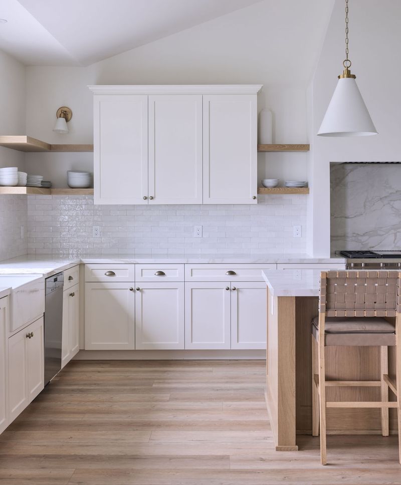 A bright, airy kitchen featuring cream shaker-style cabinetry, white subway tile backsplash, light oak open shelving, brass hardware and lighting, white quartz countertops, and light wood flooring with a woven leather bar stool at the island.