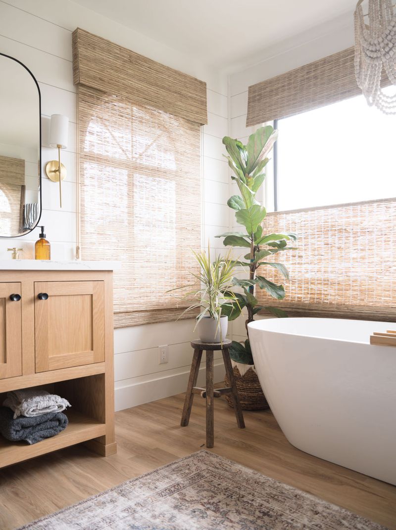 A bright, airy master bathroom featuring a light oak vanity with black hardware and open shelving, a freestanding white soaking tub, woven bamboo shades, and lush green plants on wood-look flooring.