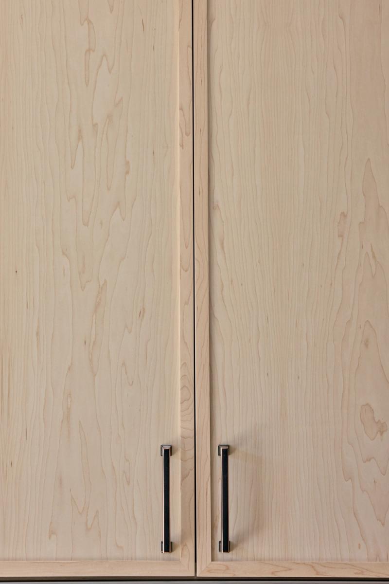 Close-up of two light maple wood cabinet doors with slim vertical bar pulls in a brushed chrome and black finish. The wood grain is pale and fine-textured.