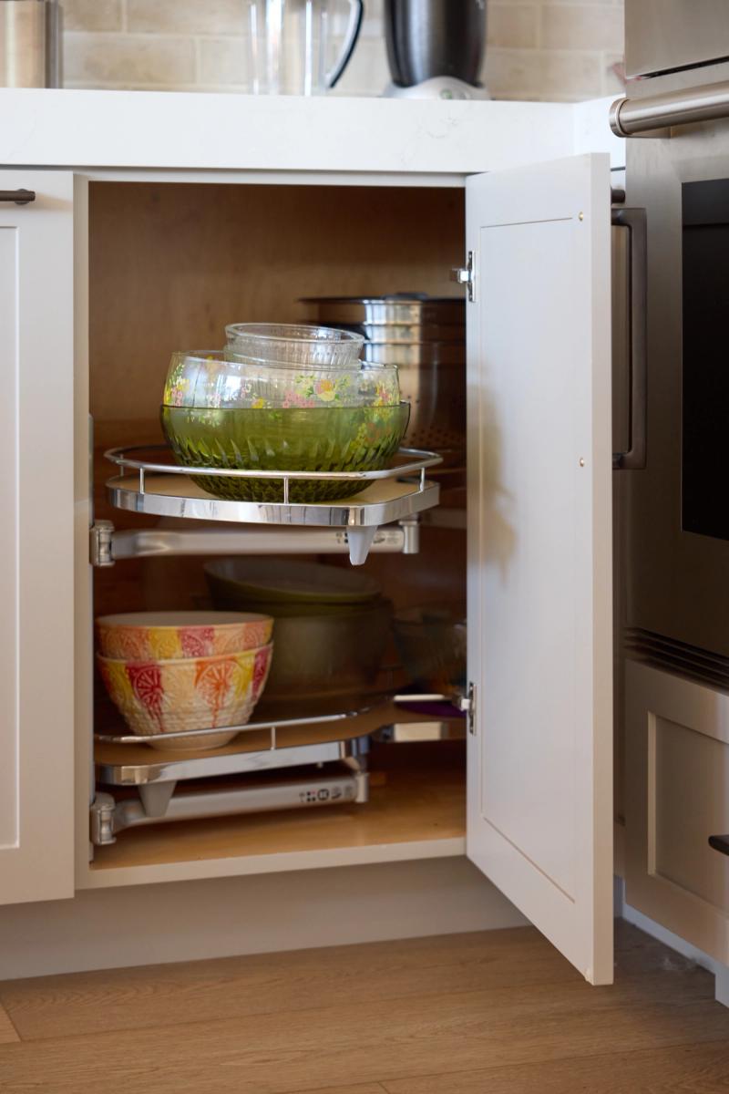 An open white shaker base corner cabinet revealing chrome lazy susan pull-out shelves holding mixing bowls and glassware, positioned next to a stainless steel oven.