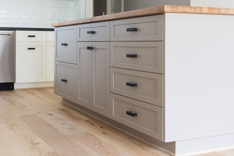 A close-up view of a kitchen island featuring greige shaker-style cabinets with matte black hardware and a warm butcher block wood countertop, set on light oak hardwood flooring. White perimeter cabinets and subway tile backsplash are visible in the background.