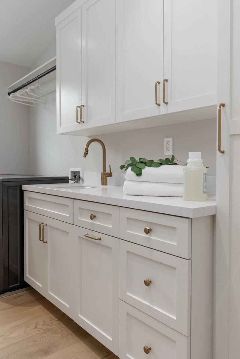 A bright, modern laundry room featuring white shaker-style cabinets with brushed brass hardware, a white quartz countertop, gold gooseneck faucet, and neatly folded white towels with a eucalyptus plant.