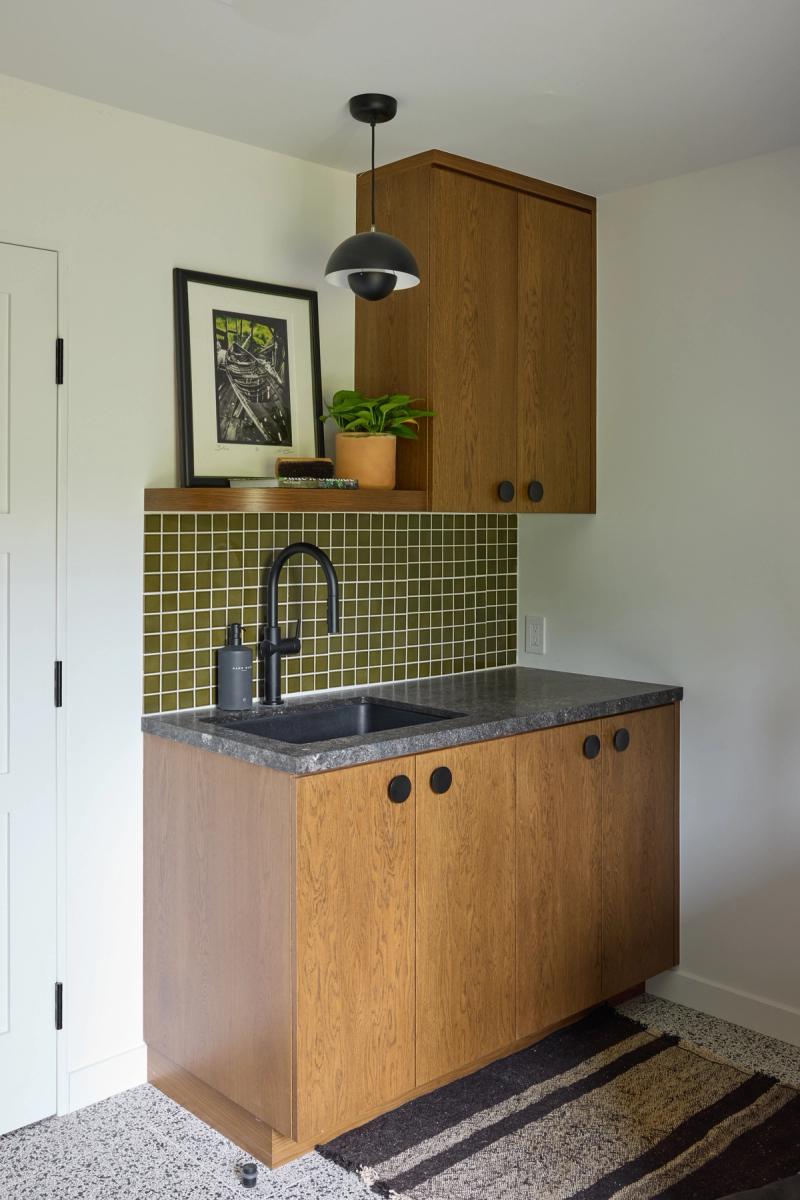 A compact wet bar or laundry sink area featuring warm oak cabinetry with black circular hardware, a dark granite countertop, olive green square tile backsplash, and a matte black gooseneck faucet, topped with a floating shelf displaying art and a potted plant.
